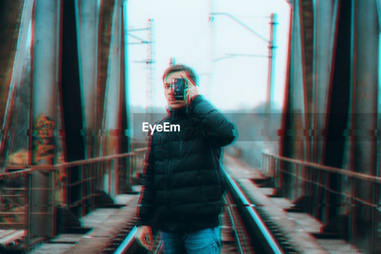 PORTRAIT OF YOUNG MAN PHOTOGRAPHING THROUGH RAILING