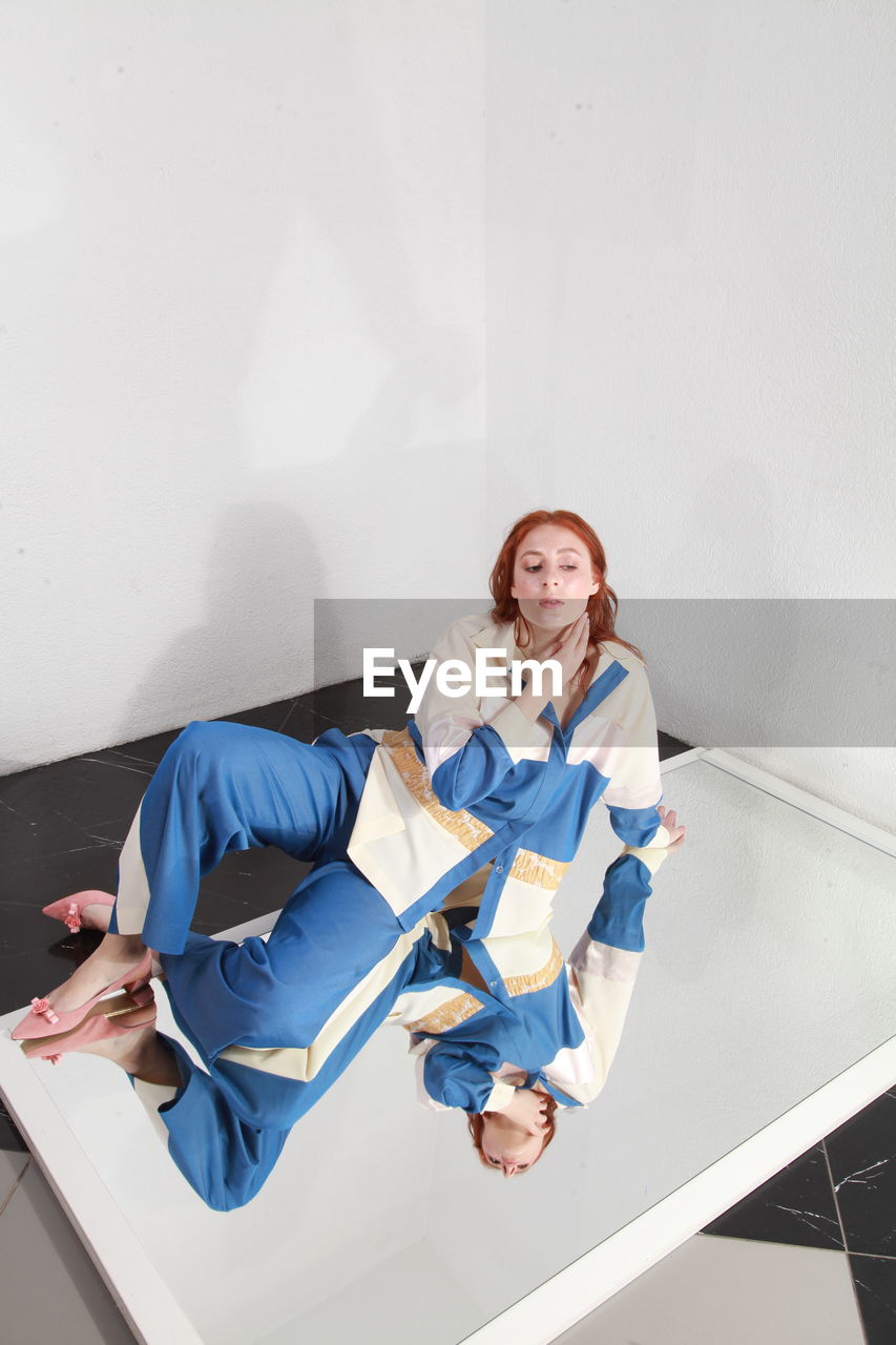 Fashion portrait of ginger woman in fashionable stylish summer suit sitting on mirror, studio