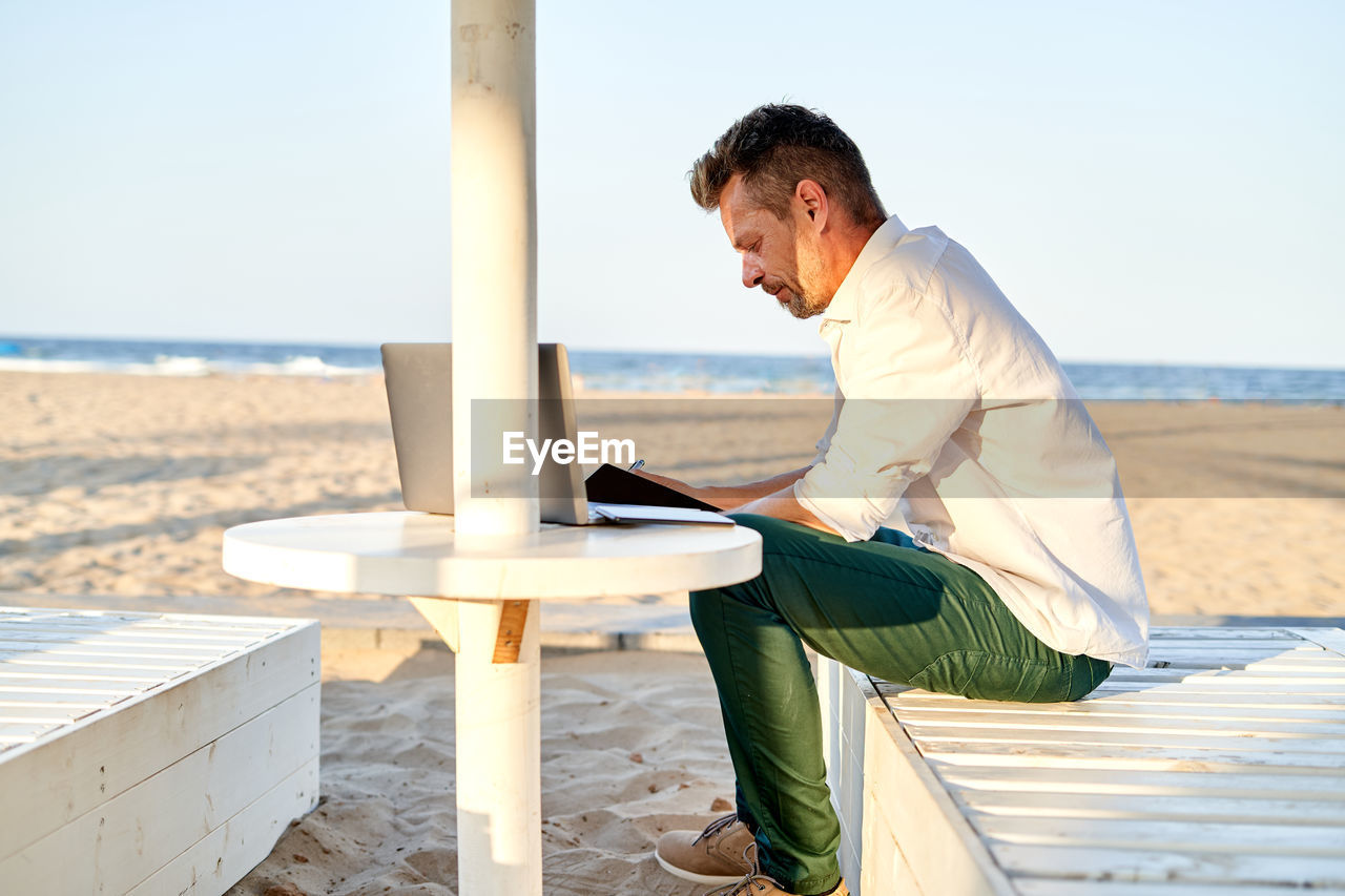 Side view of focused male entrepreneur in smart casual clothes sitting on lounger and writing in notebook while working on remote project on sandy beach near sea