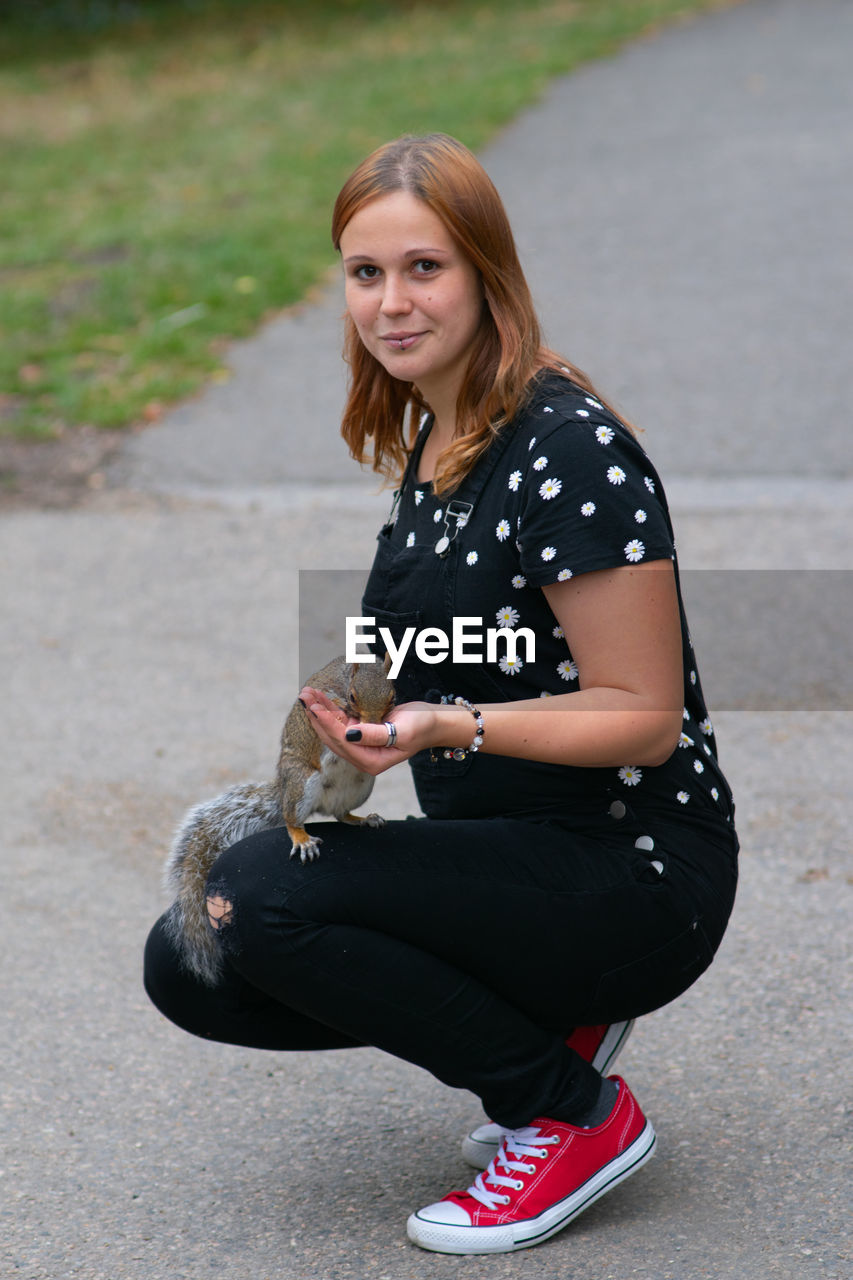 Portrait of beautiful young woman feeding squirrel while crouching on road