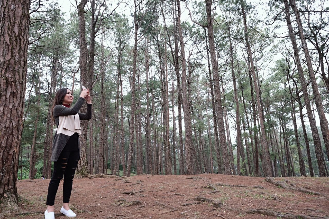 Low angle view of smiling woman photographing using phone against trees in forest