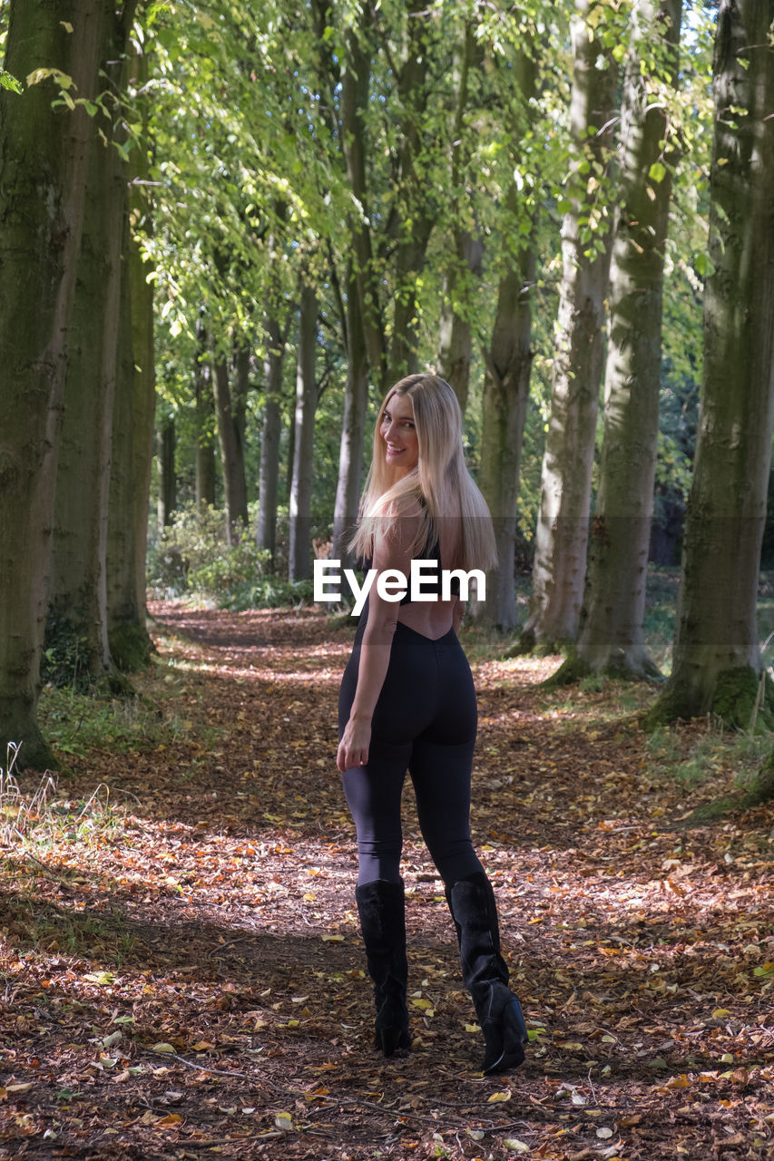 tree, one person, plant, forest, full length, adult, woodland, women, sunlight, nature, autumn, plant part, leaf, land, lifestyles, young adult, standing, leisure activity, blond hair, clothing, day, hairstyle, long hair, natural environment, outdoors, casual clothing, tree trunk, trunk, beauty in nature, tranquility, female, looking, growth, footwear