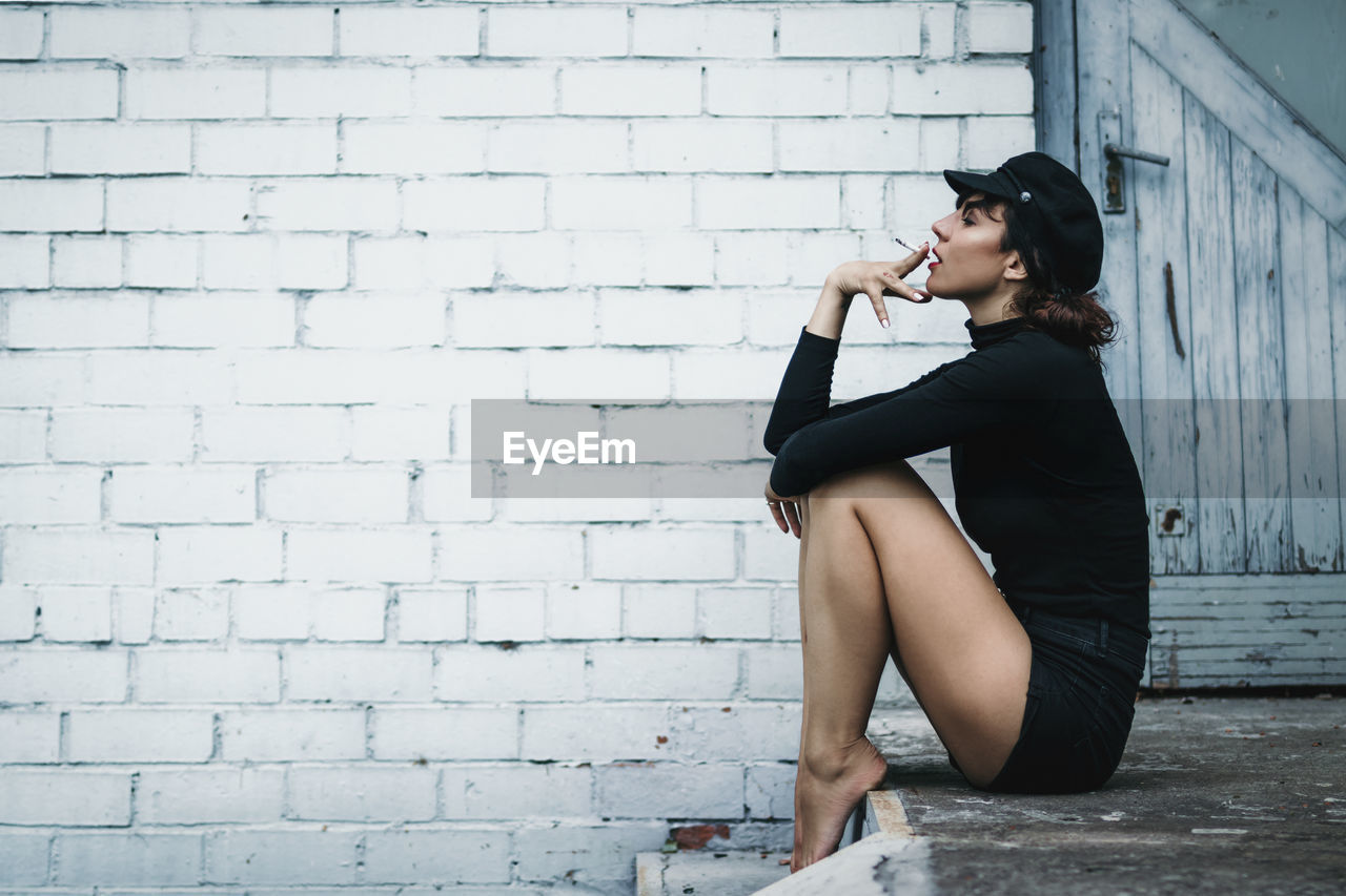 Side view of mid adult woman smoking while sitting against brick wall