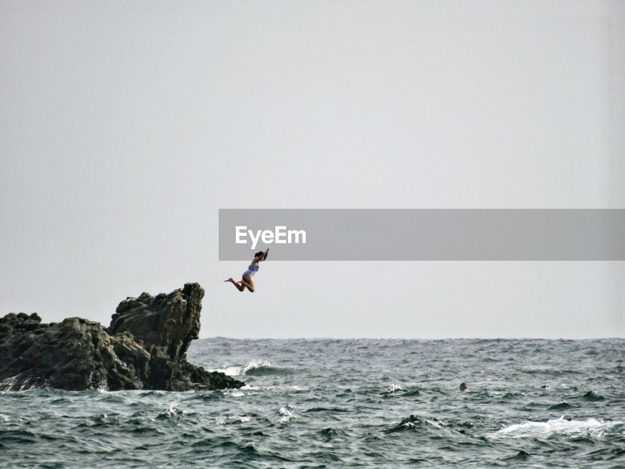 Teenage girl jumping from rock into sea against sky