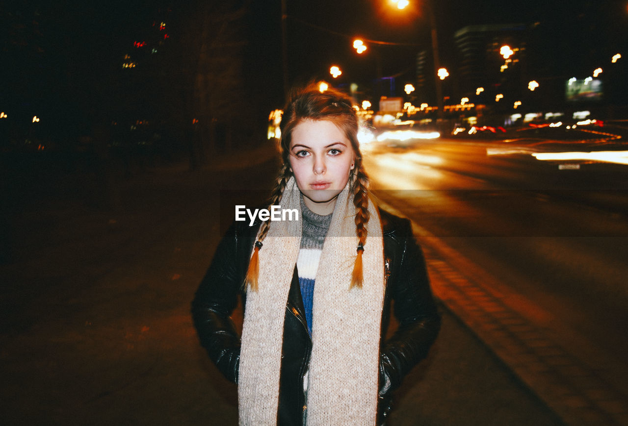 Portrait of young woman standing against light trails on road at night