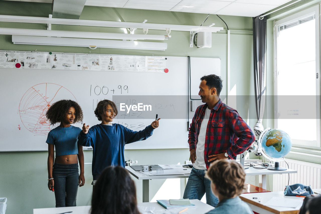 Professor with students explaining mathematics equations to pupils in classroom at school