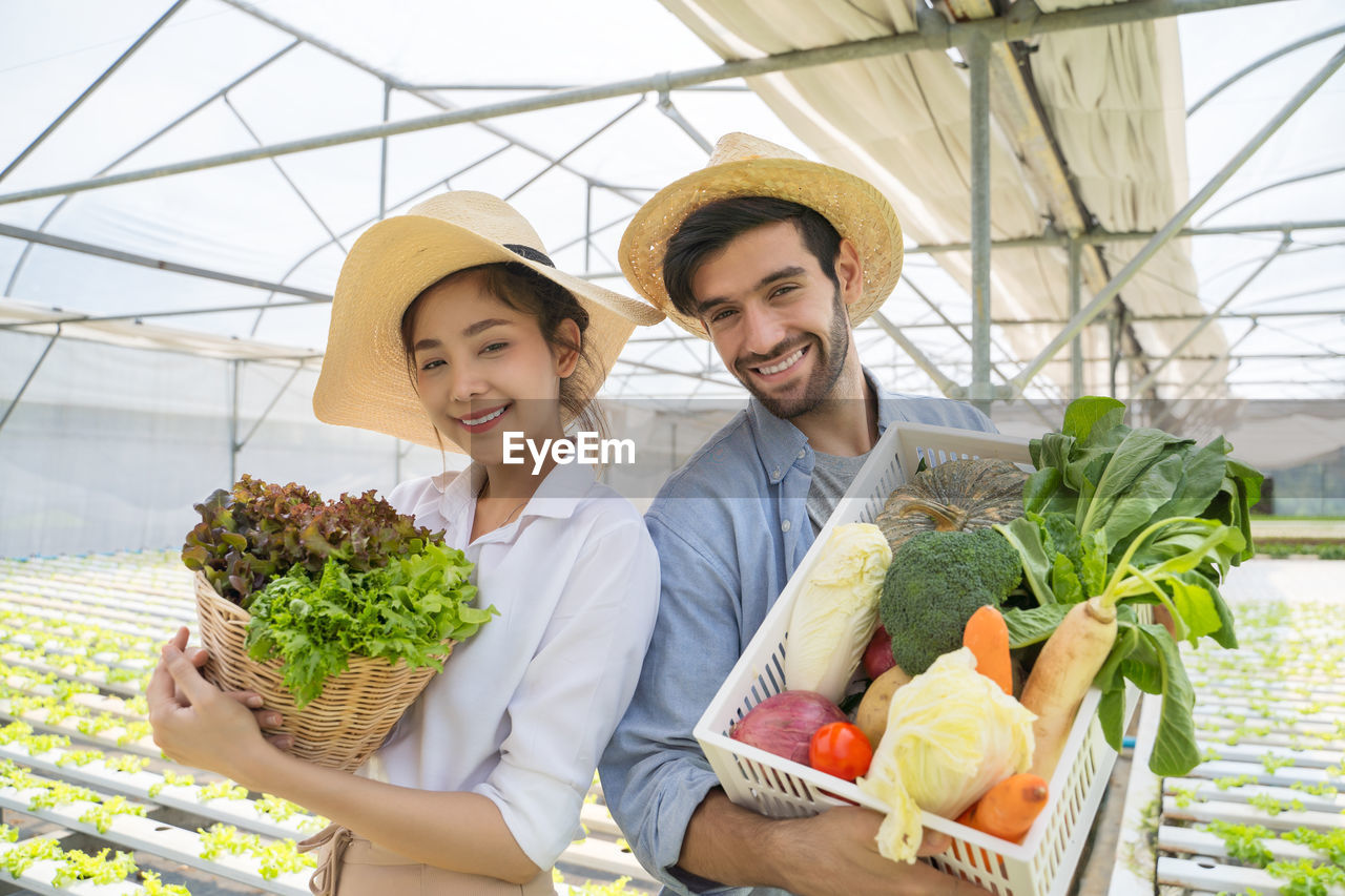 Portrait of smiling man and woman holding basket with vegetable standing at vegetable farm