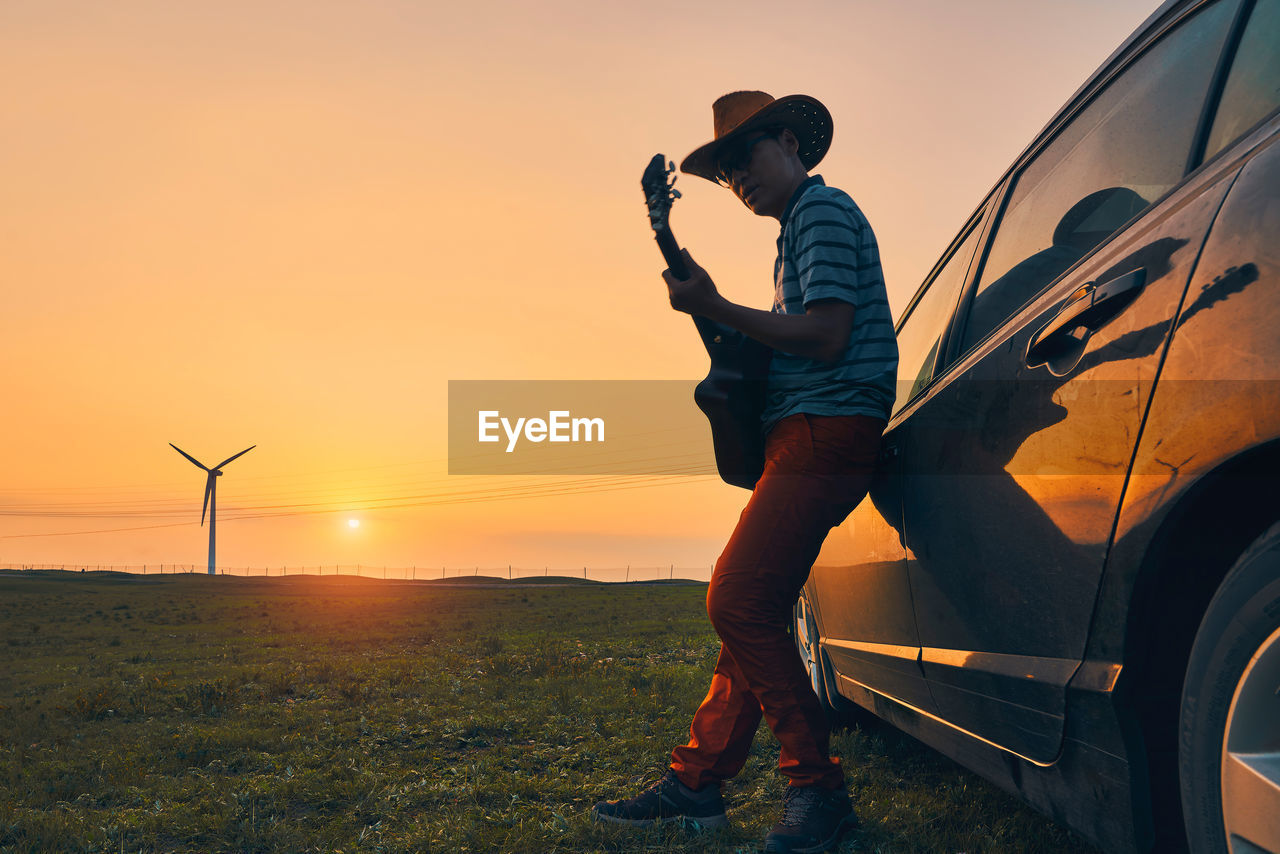 Mature man playing guitar by car on field during sunset