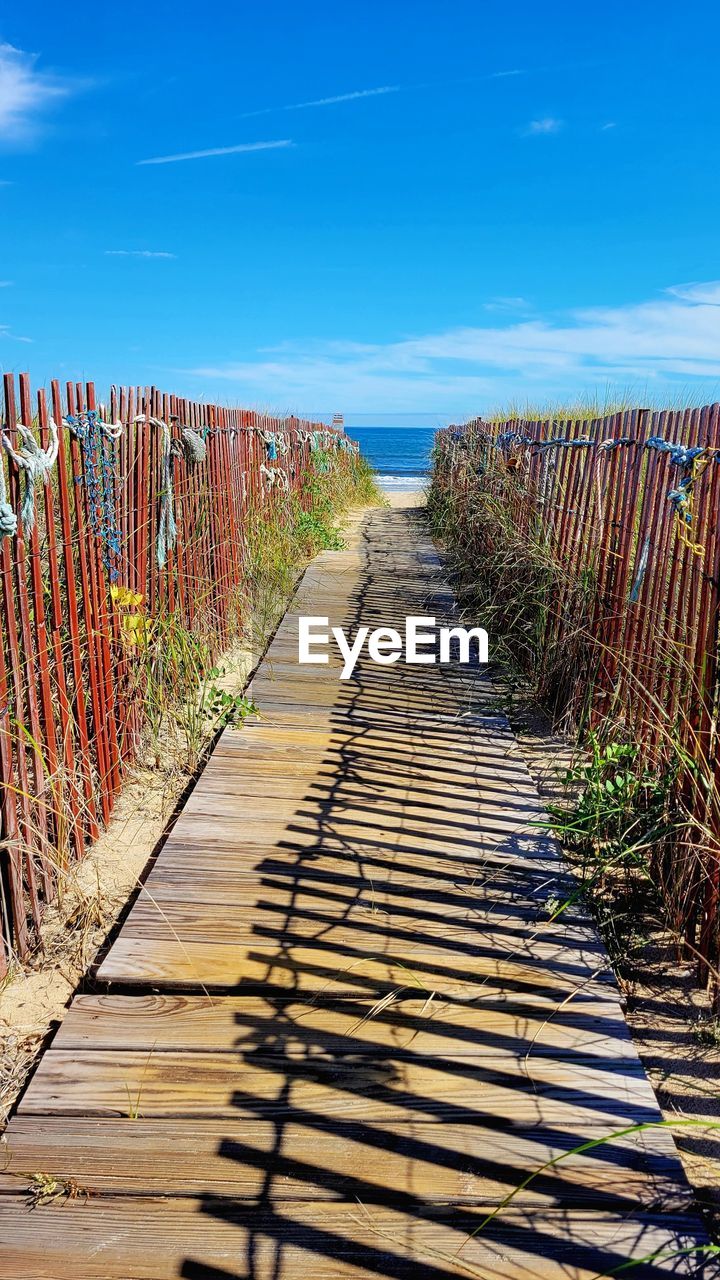 walkway, sky, nature, footpath, the way forward, boardwalk, land, no people, sunlight, blue, diminishing perspective, day, wood, scenics - nature, landscape, tranquility, plant, beauty in nature, outdoors, water, tranquil scene, sea, cloud, shadow, in a row, vanishing point, agriculture, sunny, beach, horizon, environment, travel destinations