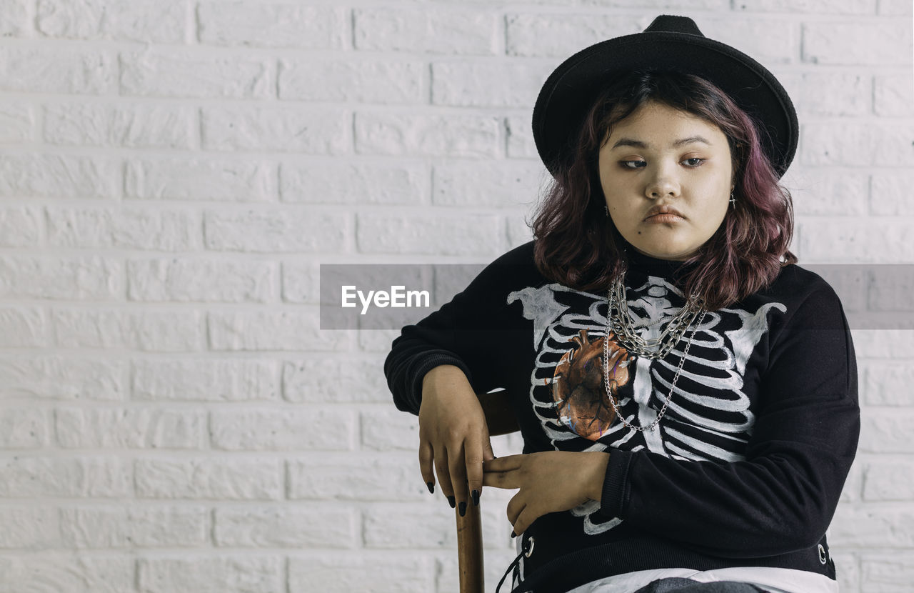 Young asian teen in costume witch sitting on a chair in front of a white brick wall,  halloween