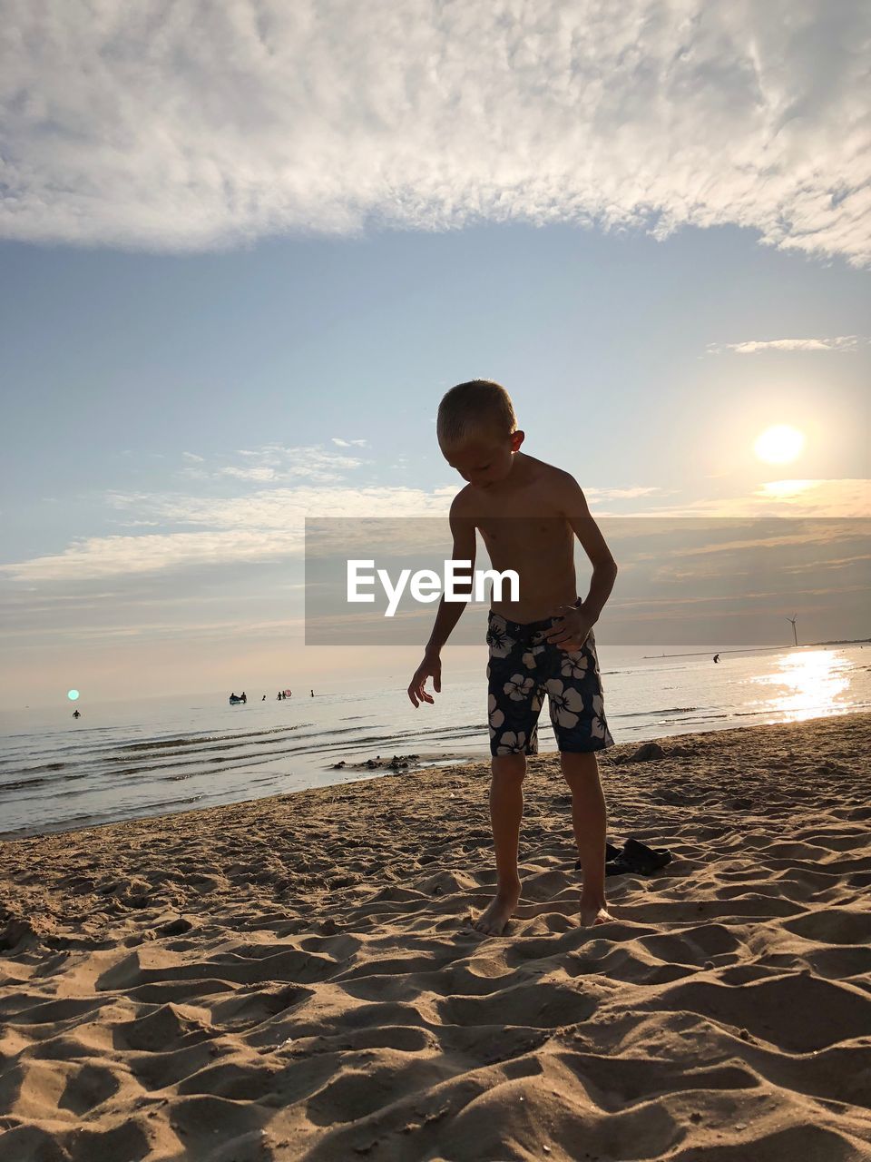 Shirtless boy standing at beach against sky during sunset
