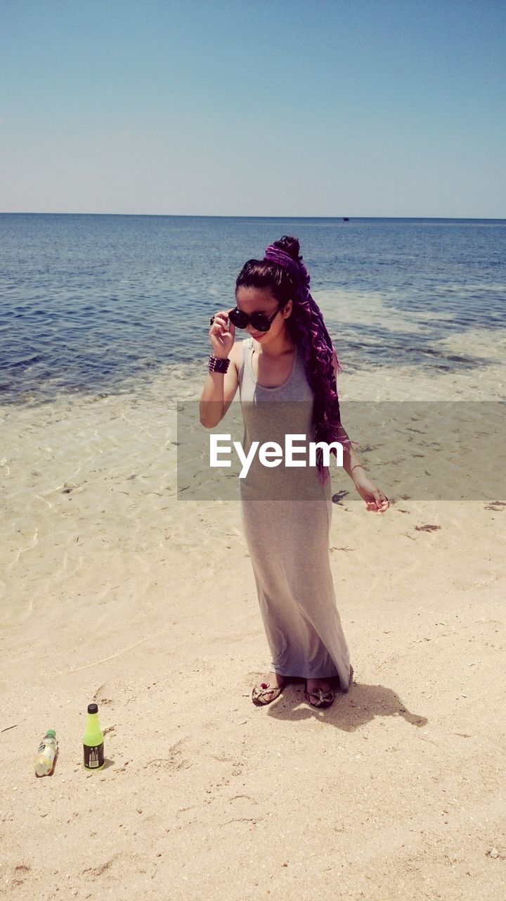 Full length of woman in sunglasses standing on beach against clear sky