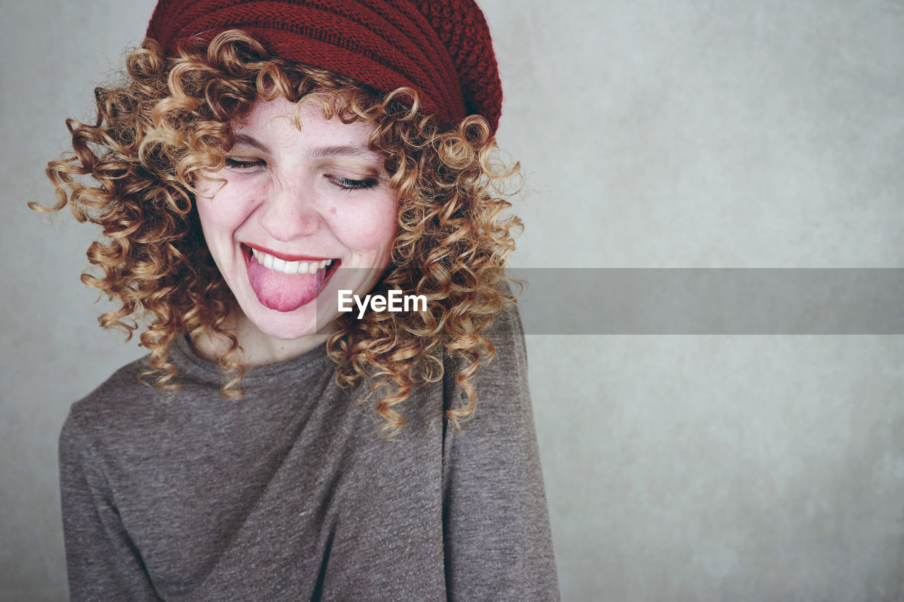 Smiling young woman sticking out tongue against wall