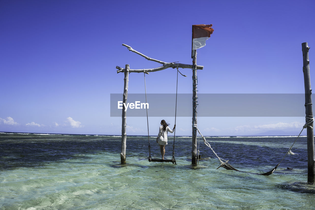 Rear view of woman standing on swing over sea against sky
