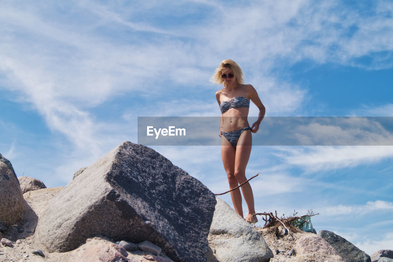 Full length of young woman in bikini standing on rock against sky
