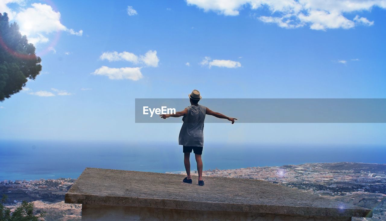 Rear view of man with arms outstretched standing on building terrace by sea against sky