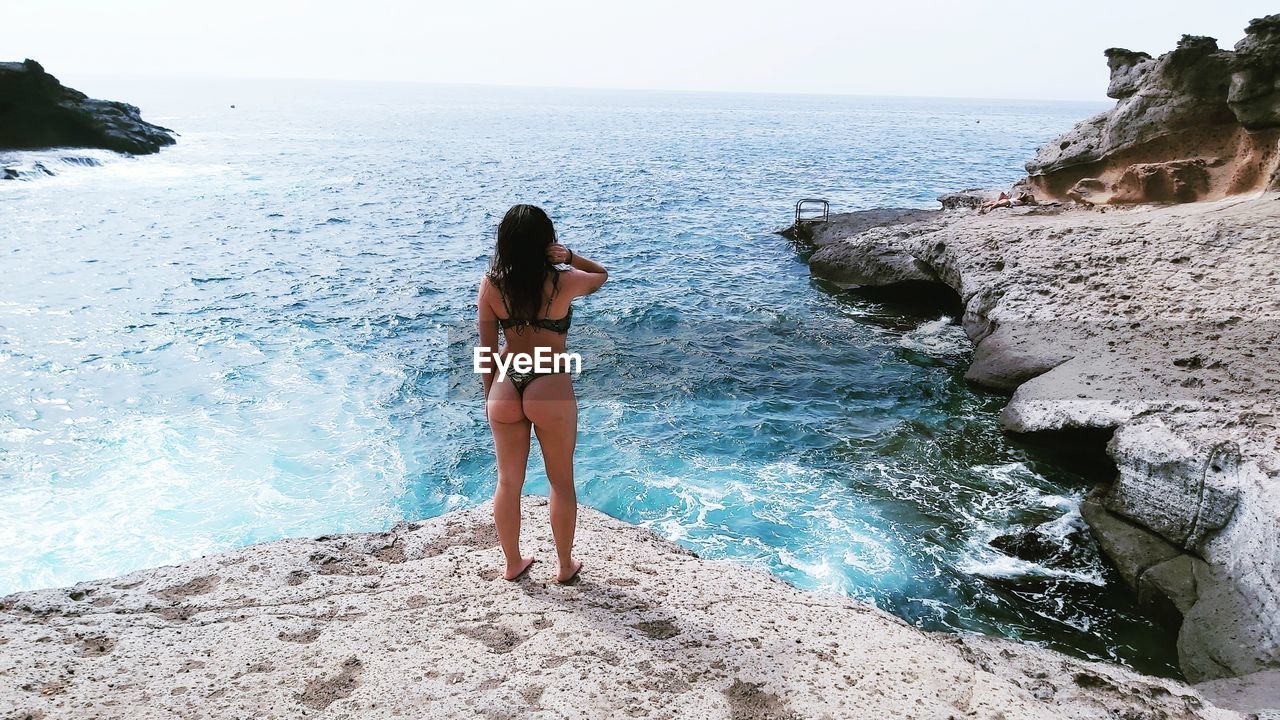 Rear view of woman wearing bikini looking at sea while standing on rock