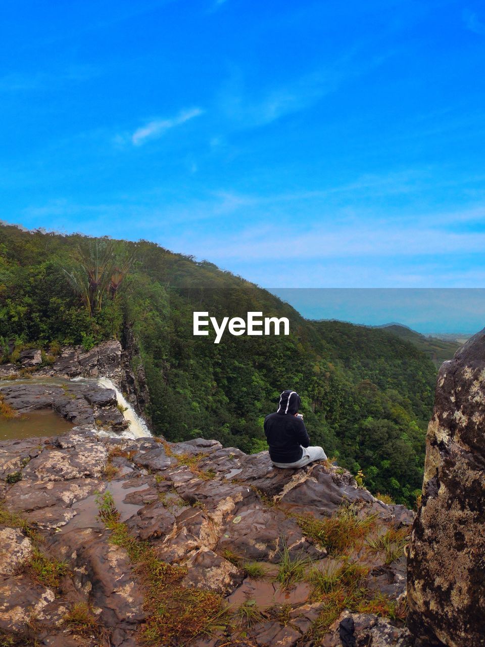 Rear view of man sitting on rock by waterfall