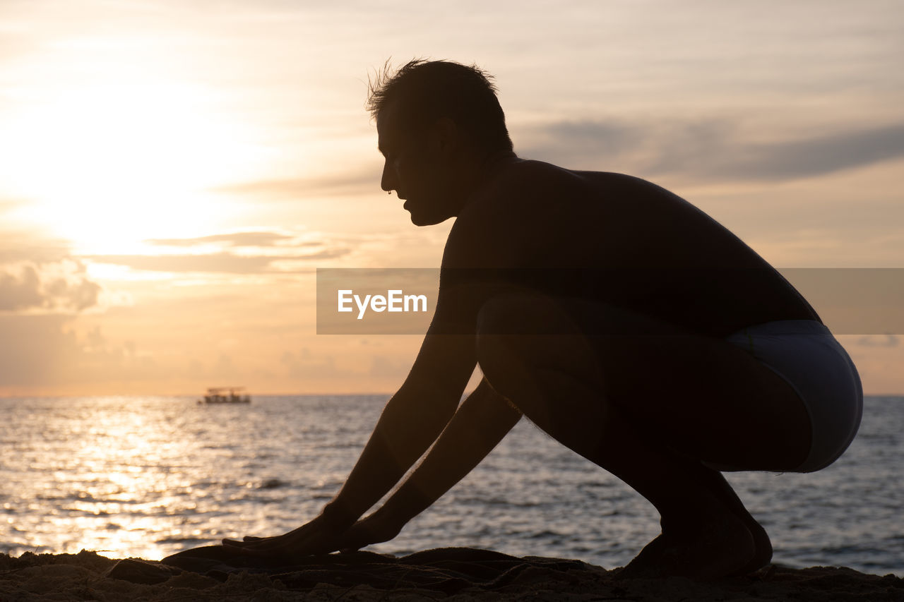 Side view of shirtless man at beach against sky during sunset