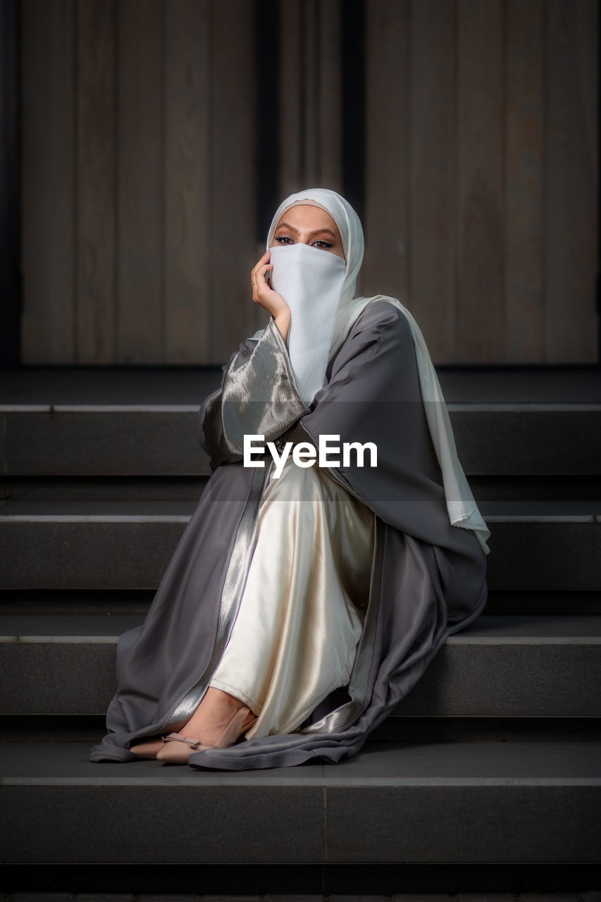 Full length image of veiled woman with white niqab sitting on the stairs and looking at the camera