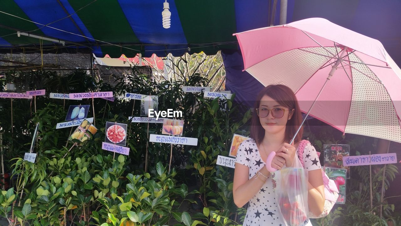 Portrait of young woman with umbrella standing against plants at farm