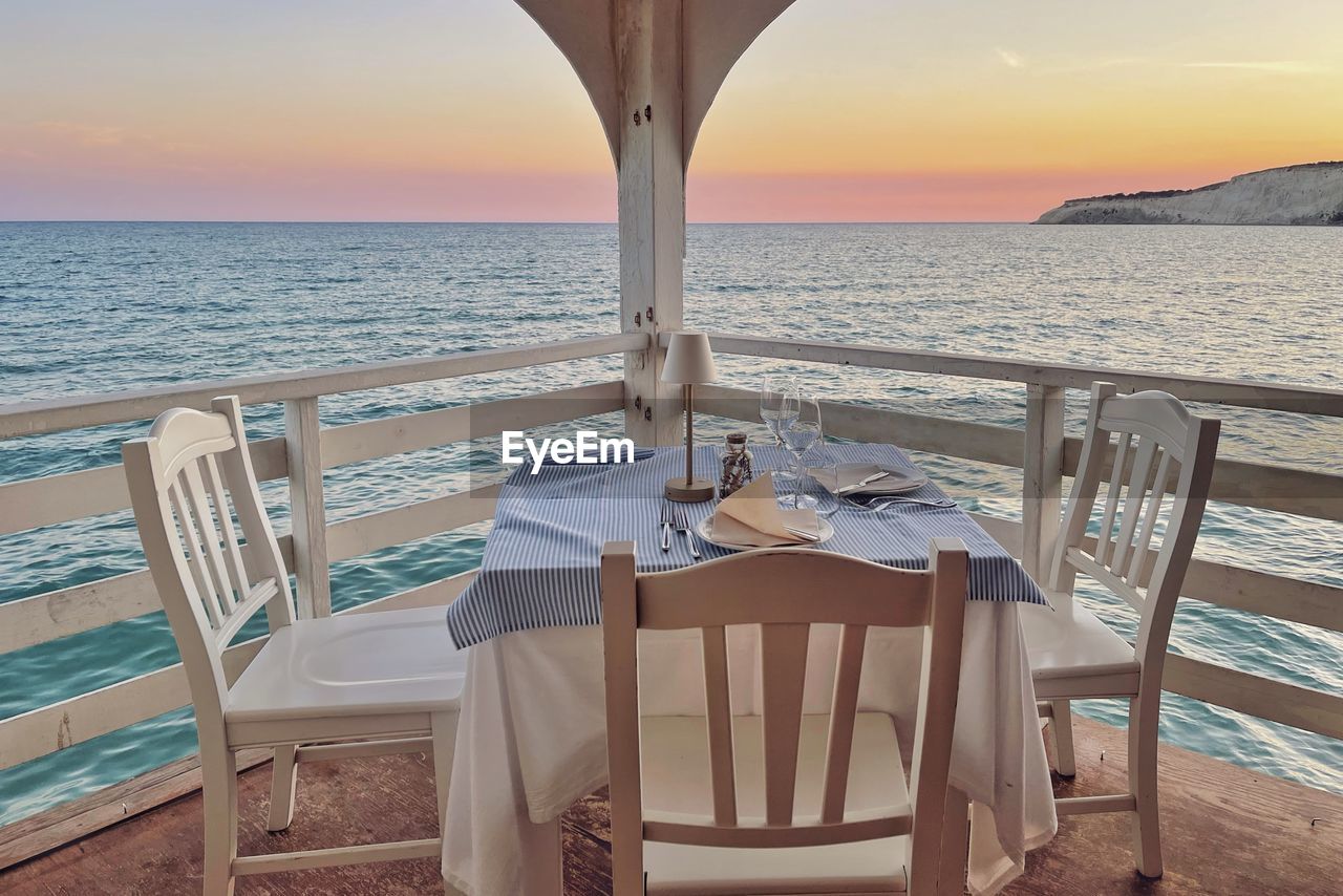 chairs and tables at beach against sky during sunset