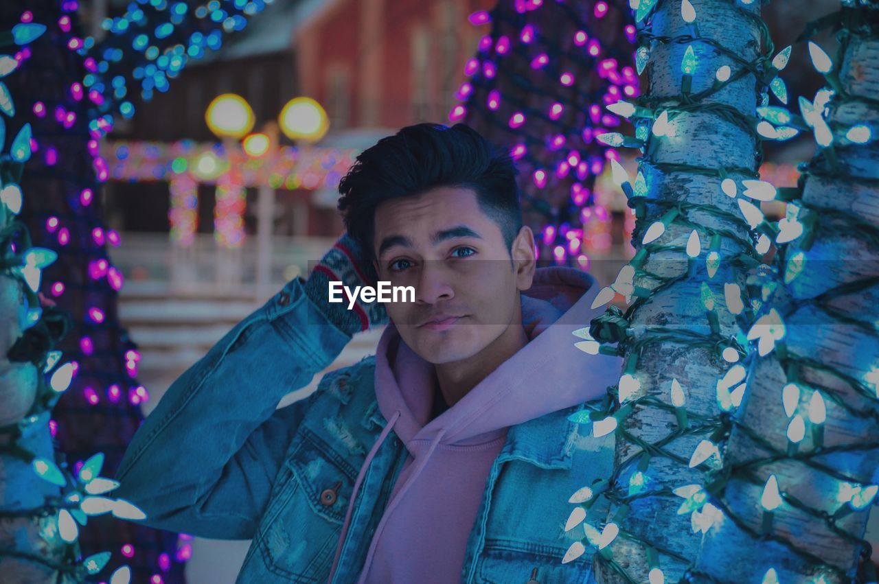 Portrait of young man standing by illuminated christmas tree at night