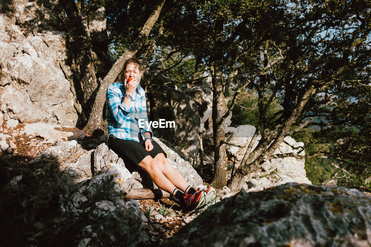 Portrait of woman eating apple while sitting on rock against trees