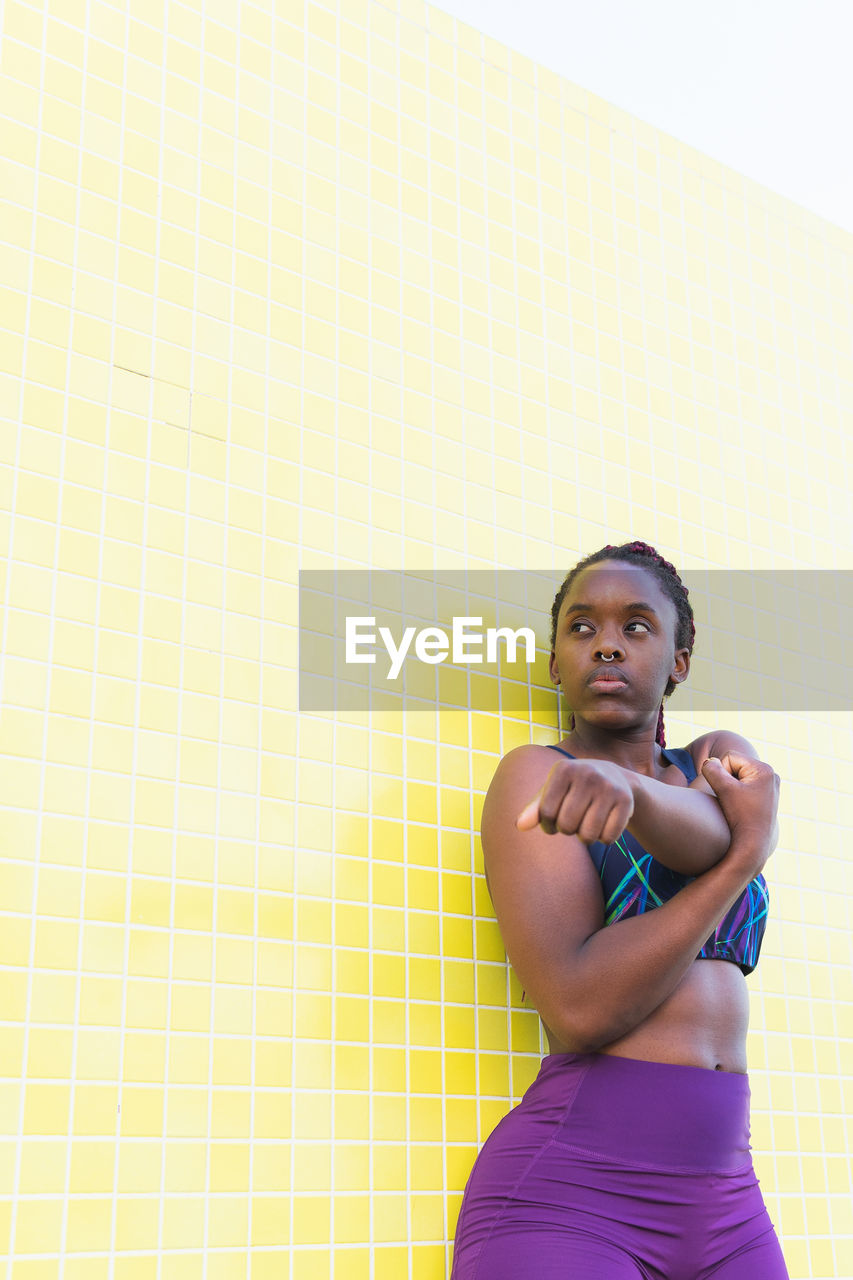 Fit black female athlete in sportswear doing stretching exercises while warming up before training against yellow tiled wall