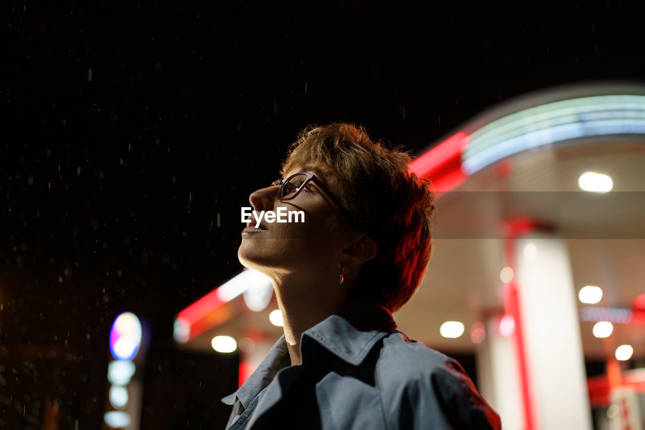 Caucasian woman in glasses is standing next to car filling station and enjoying falling snowflakes