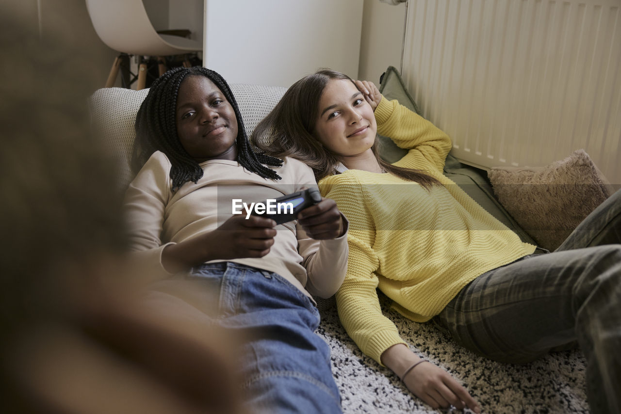 Smiling teenage girl holding game controller while lying down with female friend in bedroom