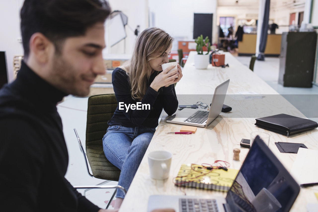 Young woman drinking coffee while sitting with colleague at desk in office