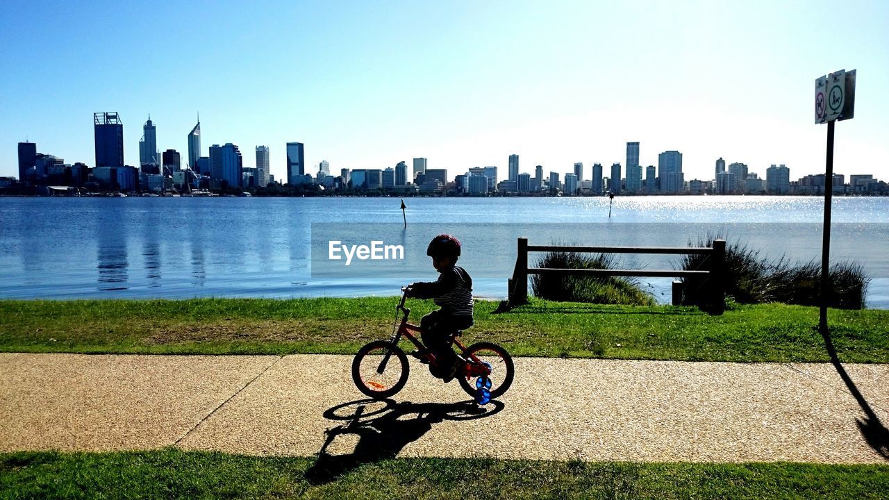 Side view of boy riding bicycle on footpath by river against buildings in city