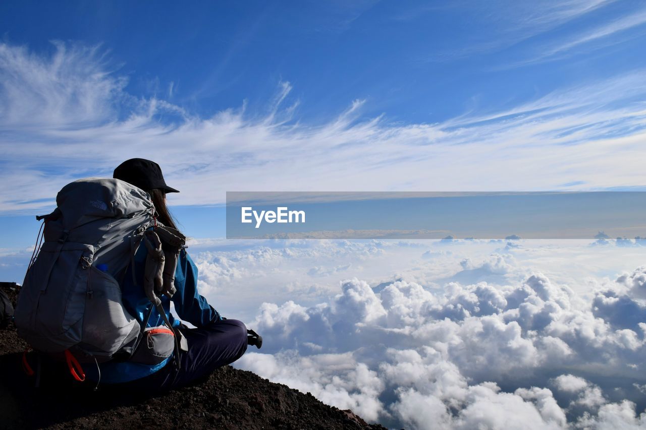 Hiker with backpack sitting on cliff against cloudy sky