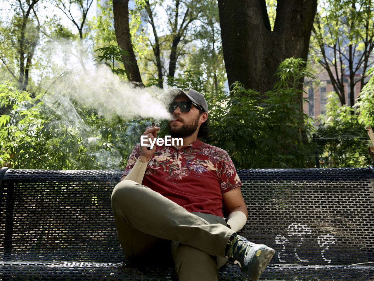 Young man sitting on table against trees smoking marijuana