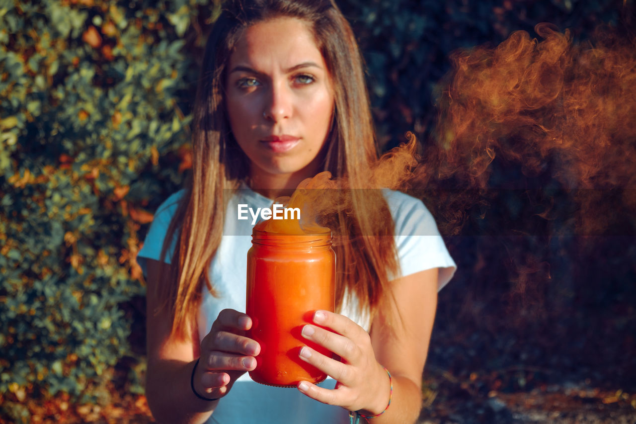 Portrait of woman holding jar with orange smoke while standing against plants
