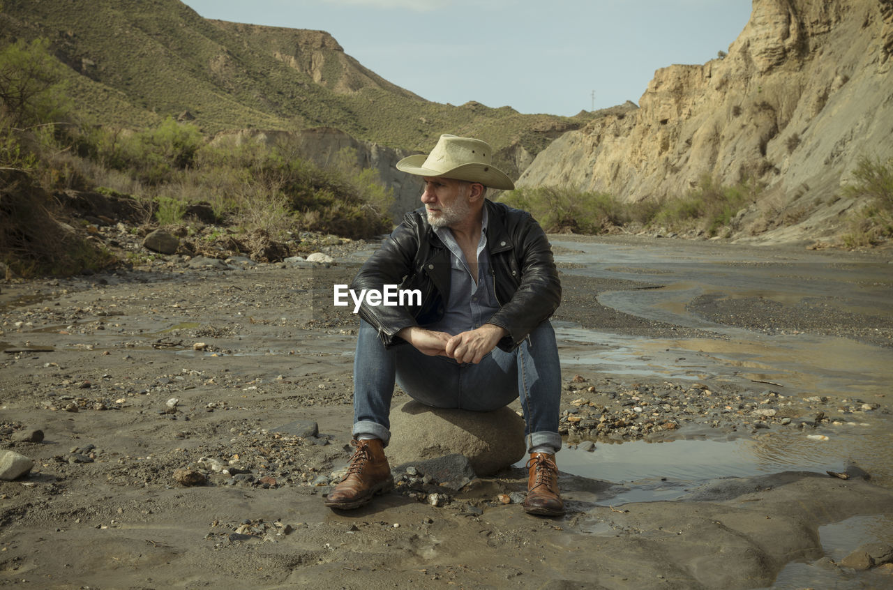 Adult man in cowboy hat sitting on rock along river flows in desert. almeria, spain