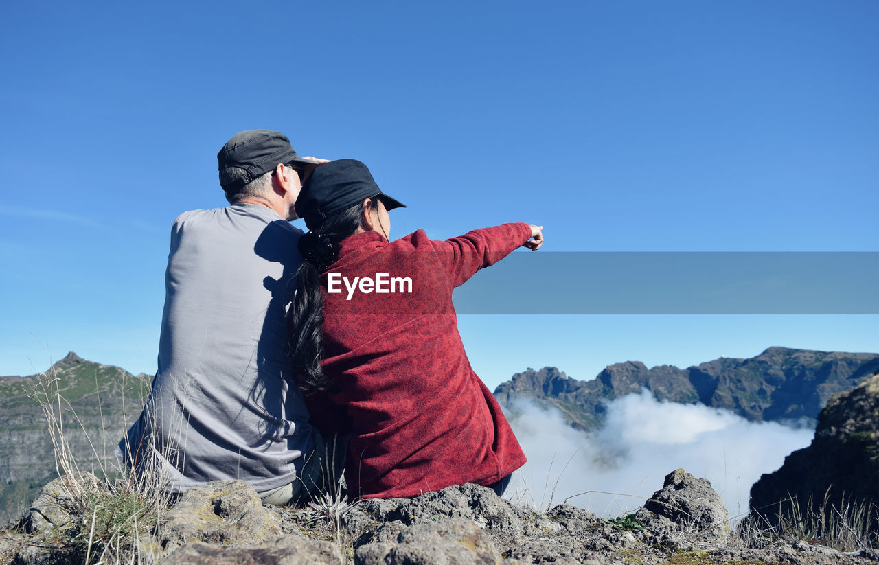 Rear view of couple sitting on mountains against clear blue sky