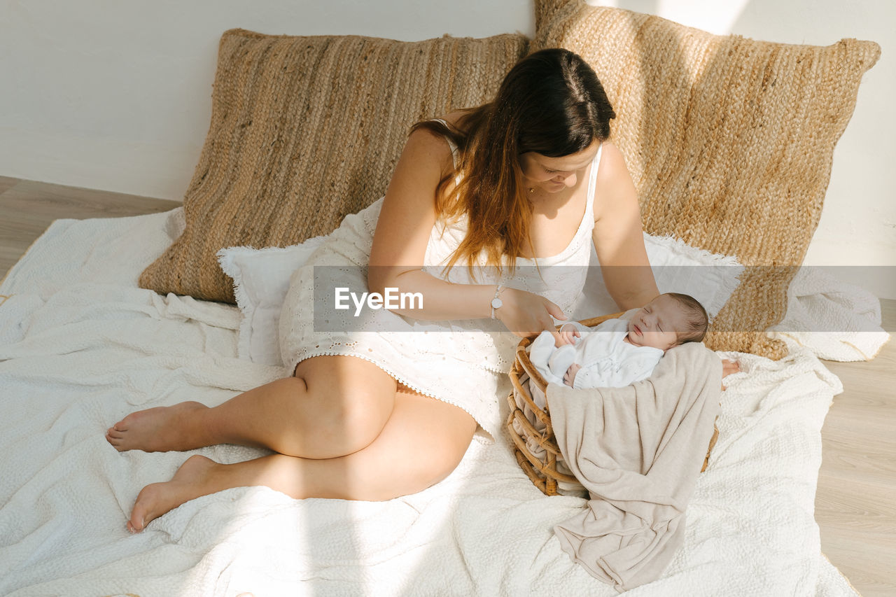 High angle of barefoot woman touching newborn baby sleeping in basket while sitting on blanket near pillows in bedroom