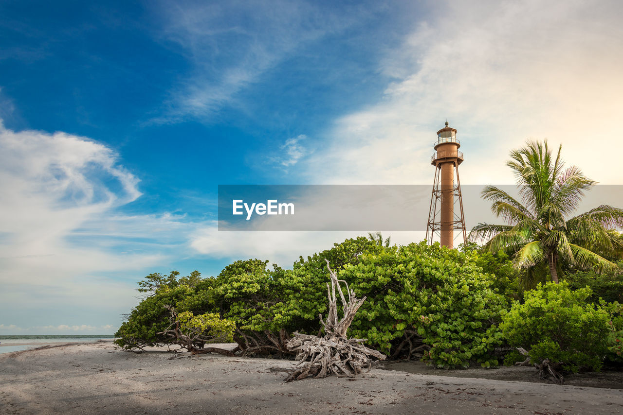 Low angle view of lighthouse amidst trees by beach against cloudy sky