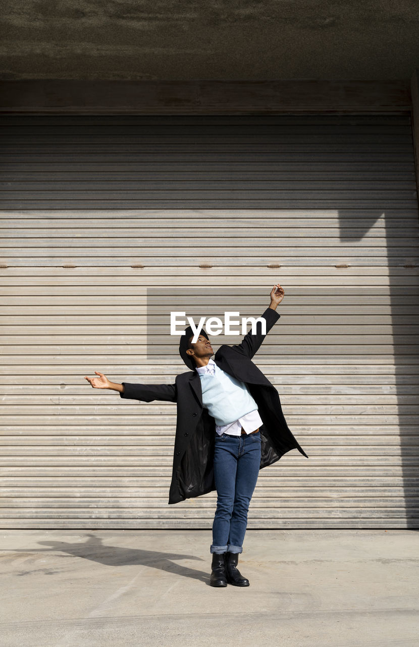 Young man wearing hat with arms outstretched dancing against shutter