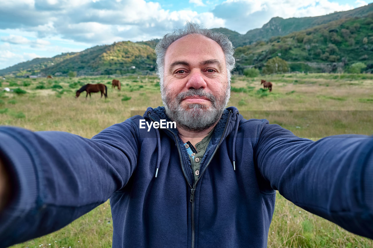 Happy, serious mature bearded man making a selfie on meadow with grazing horses.