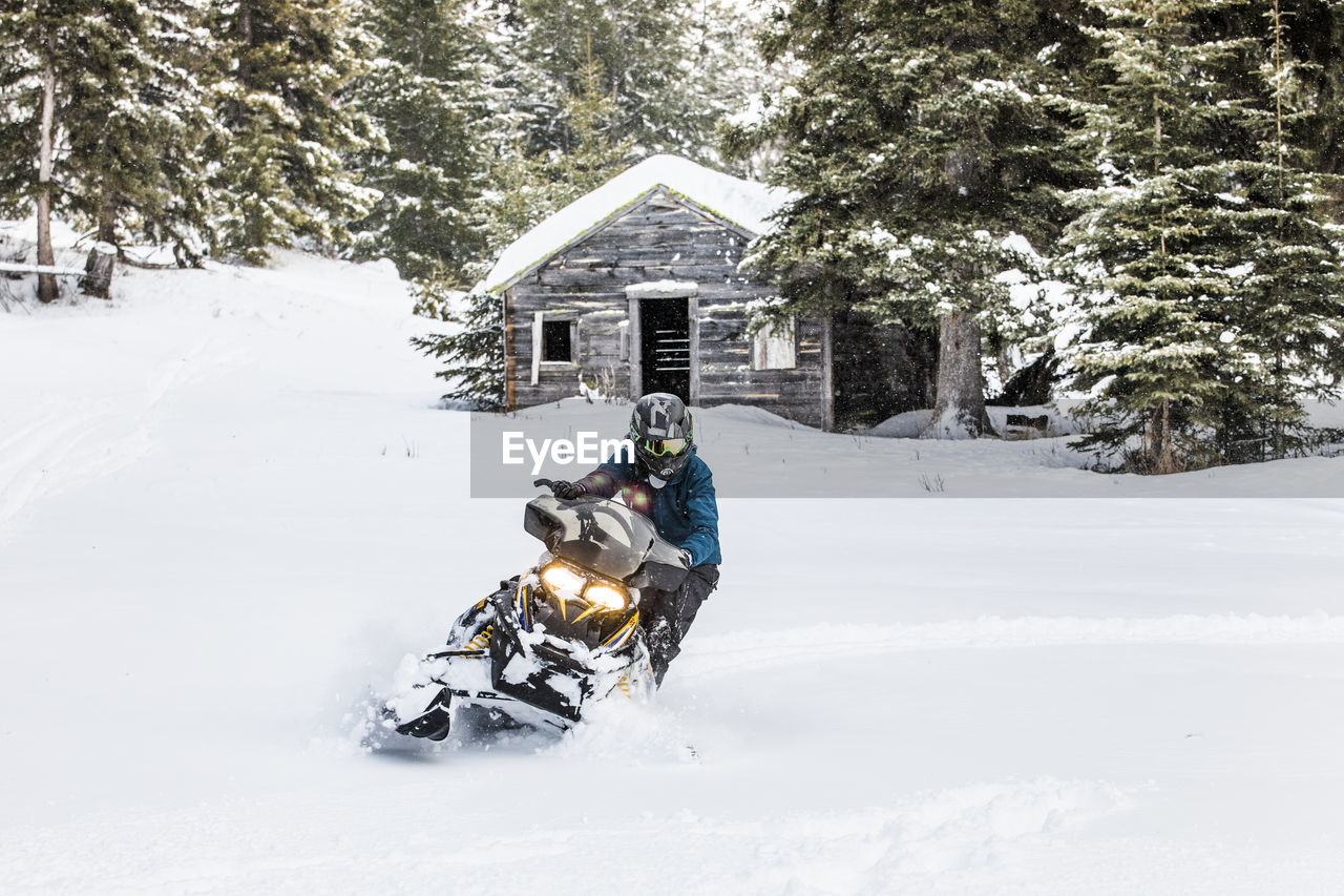 Confident man on snowmobile with old cabin behind.