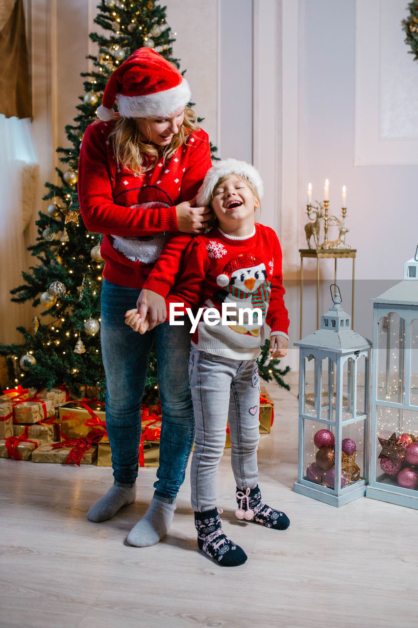 Happy mom and laughing daughter in christmas sweaters stand by the christmas tree and have fun