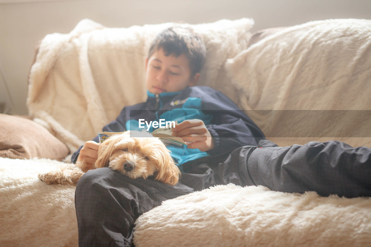 Boy with book sleeping while sitting by dog on sofa at home