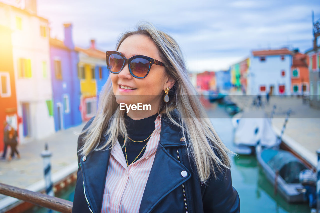 portrait of young woman wearing sunglasses while standing against buildings