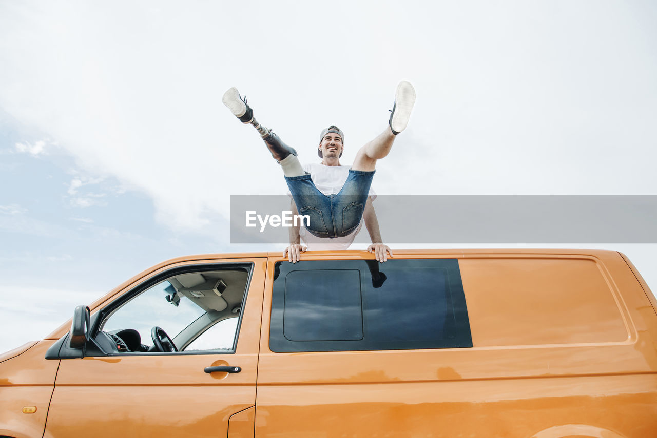 Happy young man with leg prosthesis balancing on roof of camper van