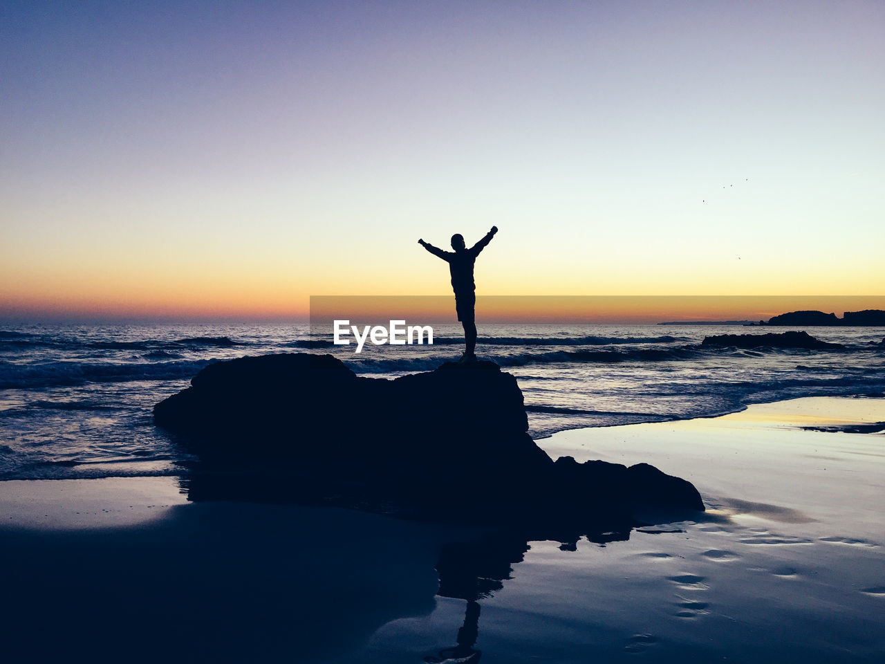 Silhouette person with arms outstretched standing on rock at beach during sunset