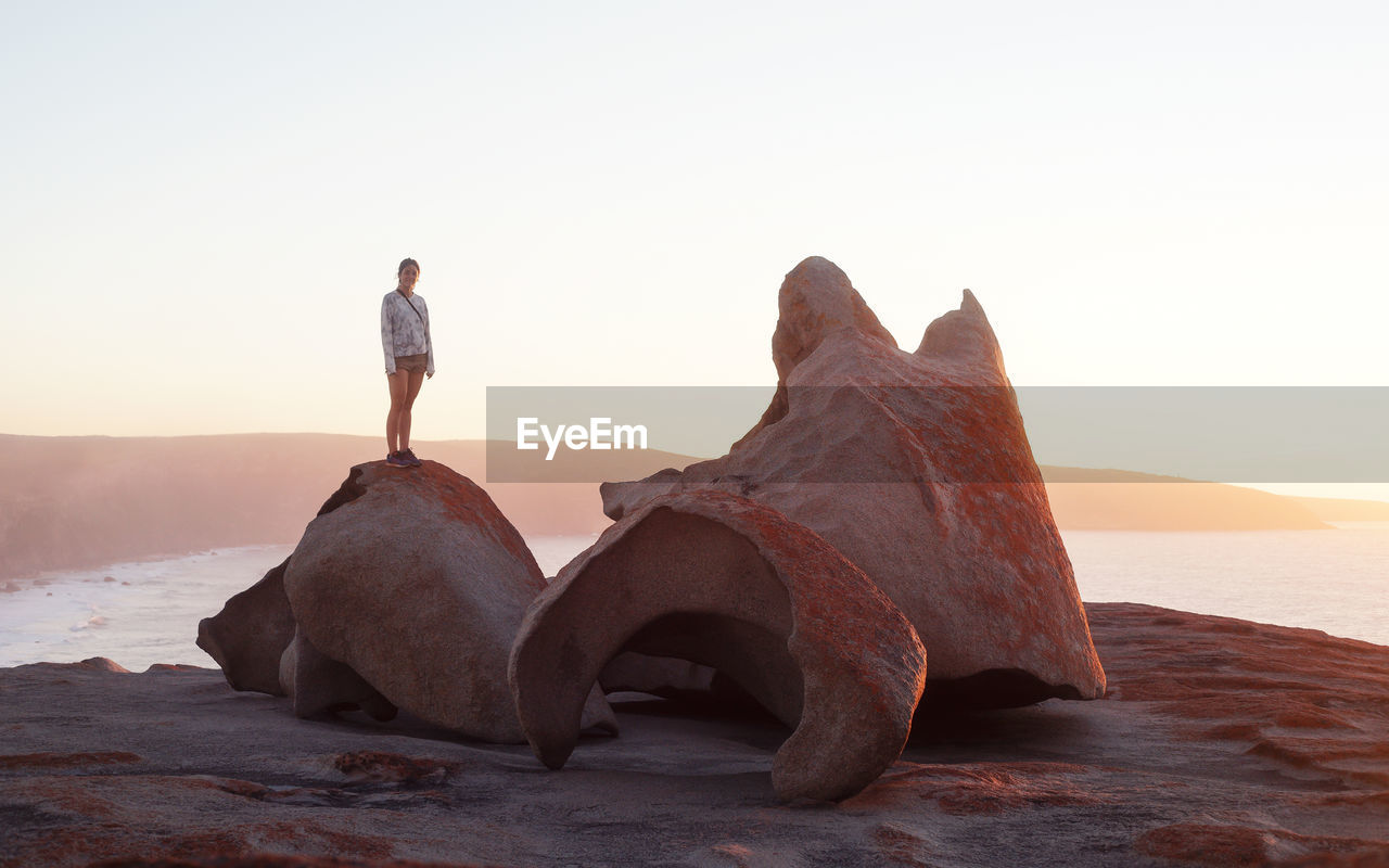 Woman standing on rock at beach against clear sky during sunset