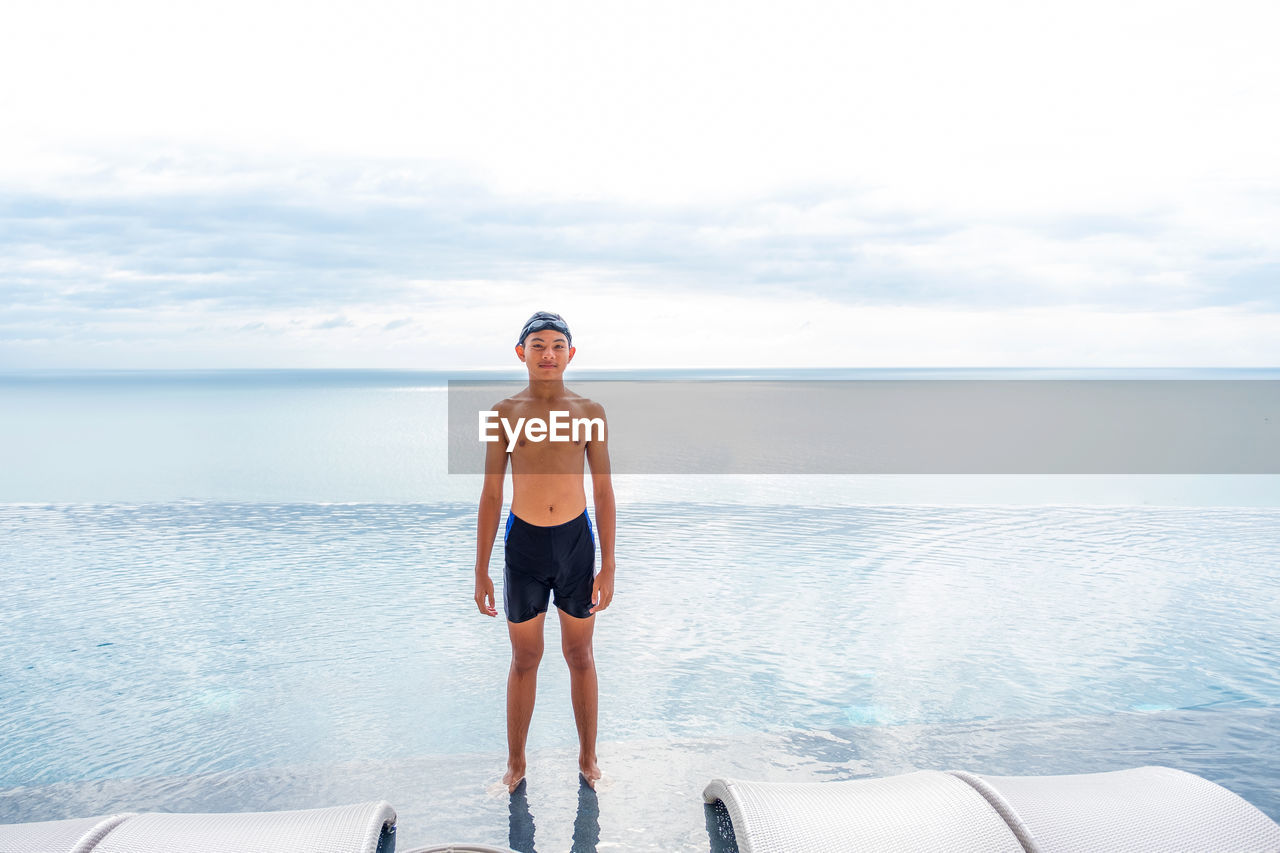 Portrait of shirtless boy standing by swimming pool against sky